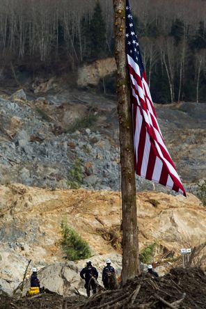 On April 2, FEMA workers stand beneath an American flag flying at half staff in the debris field of the Highway 530 mudslide, west of Darrington. The large flag, raised on the third day after the mudslide, replaces an older flag found in the debris field and raised at half staff by the Oso Fire Department on the second day.